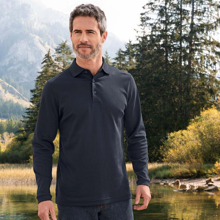 Mann in schwarzem Pima-Interlock-Poloshirt, steht in einer natürlichen Umgebung mit Bergen im Hintergrund.