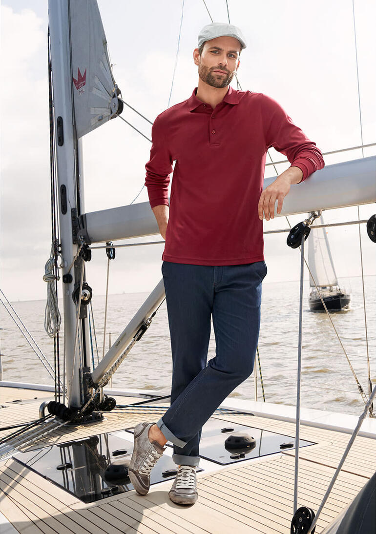 Seiden-Jeans Mann in rotem Pima Interlock Polo Shirt steht auf einem Boot, lächelt.