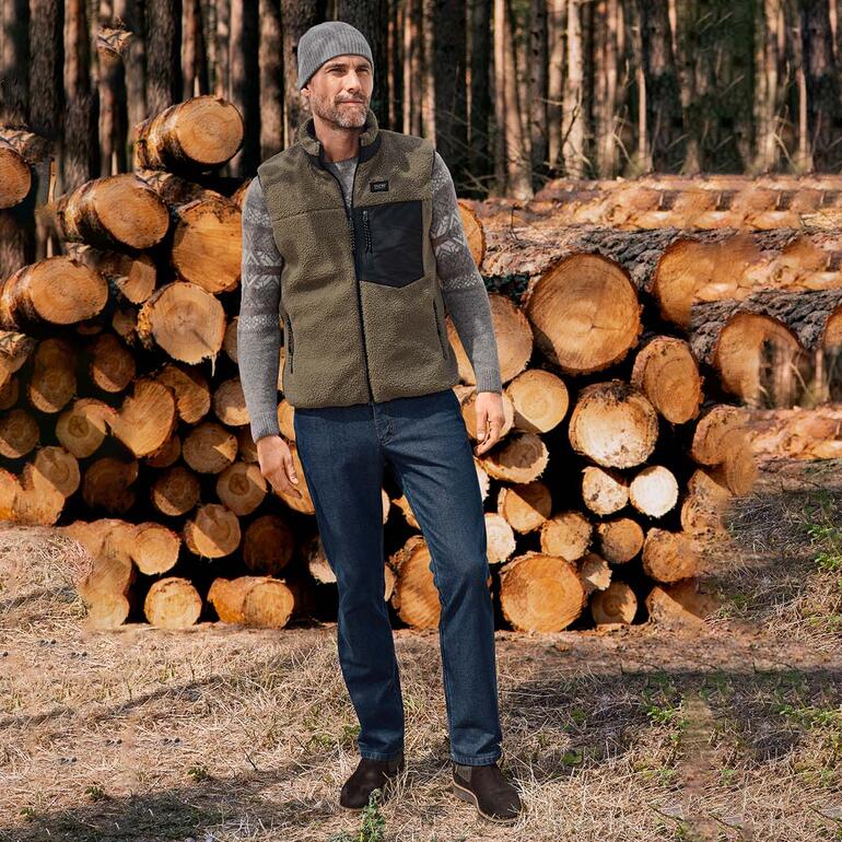 Mann trägt einen Baby-Alpaka Norwegerpullover, steht vor einem Holzstapel im Wald.