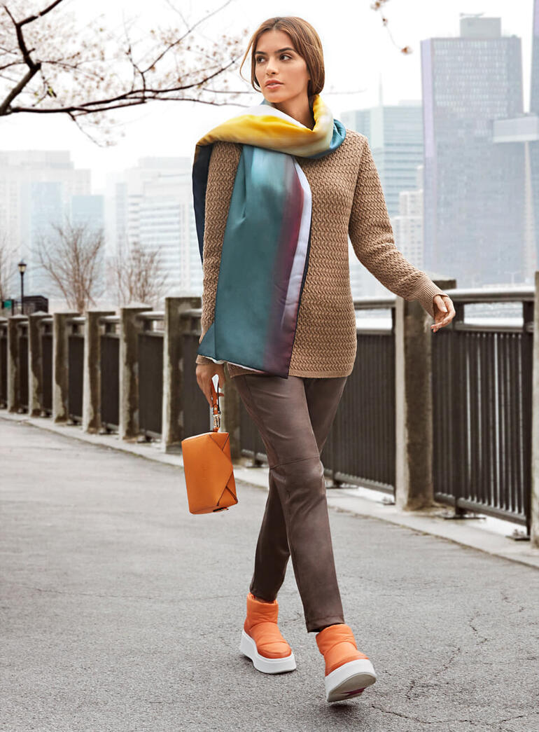 Frau in Kamelhaar-Pullover wandert durch eine Stadt mit Uferpromenade.