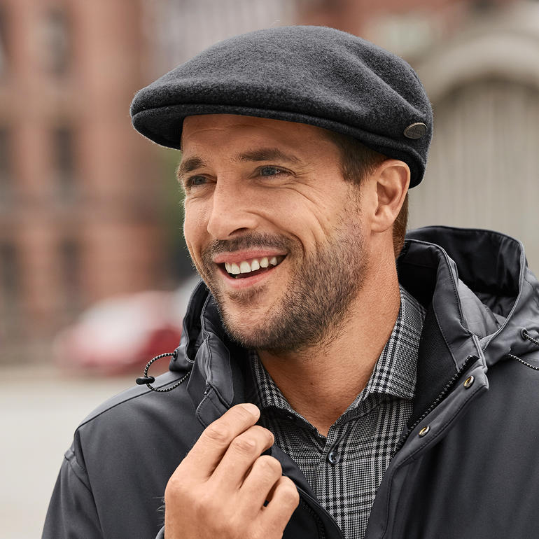 Mann mit Kangol-Schiebermütze und Ohrenwärmern, lächelt in urbaner Umgebung.