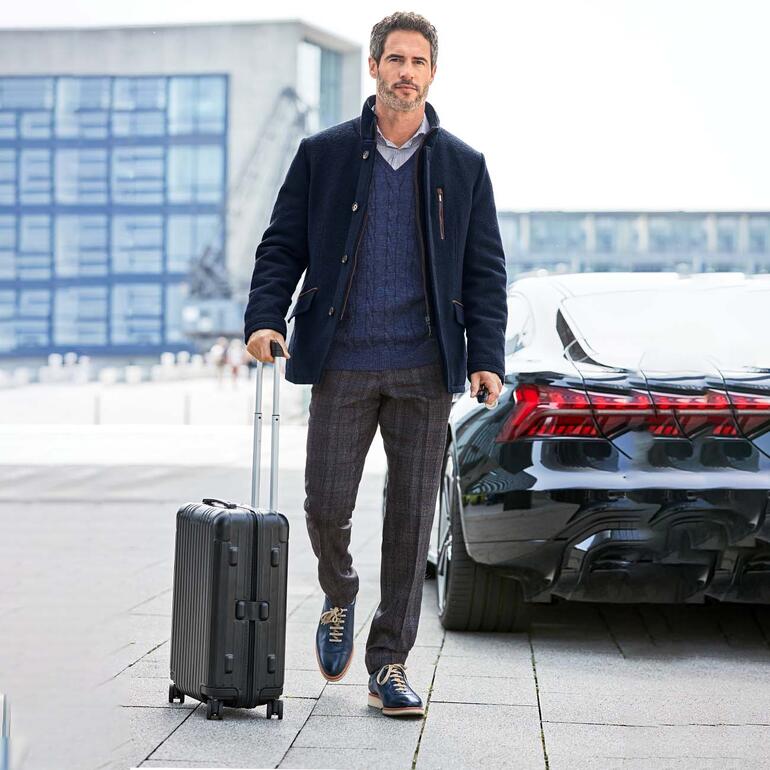 Mann mit Wollwalk-Sakko am Flughafen, neben einem schwarzen Auto.