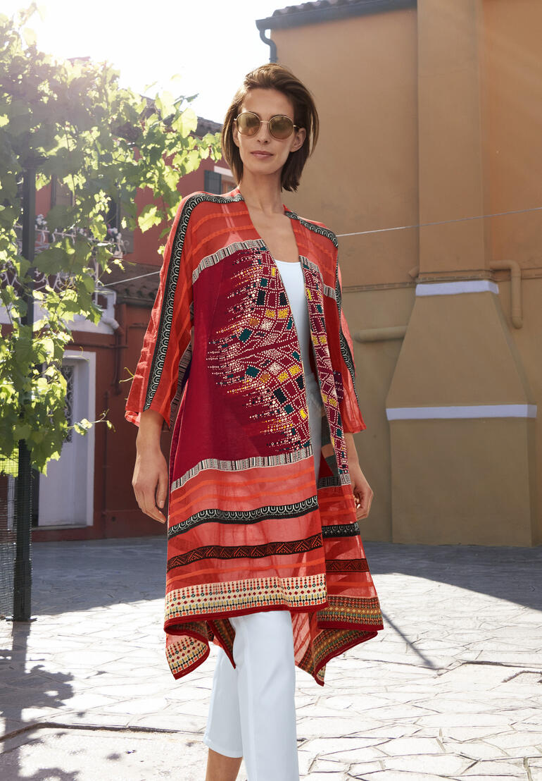 Frau in Rot mit IVKO Kimono-Strickjacke, steht im Sonnenlicht auf einem Platz