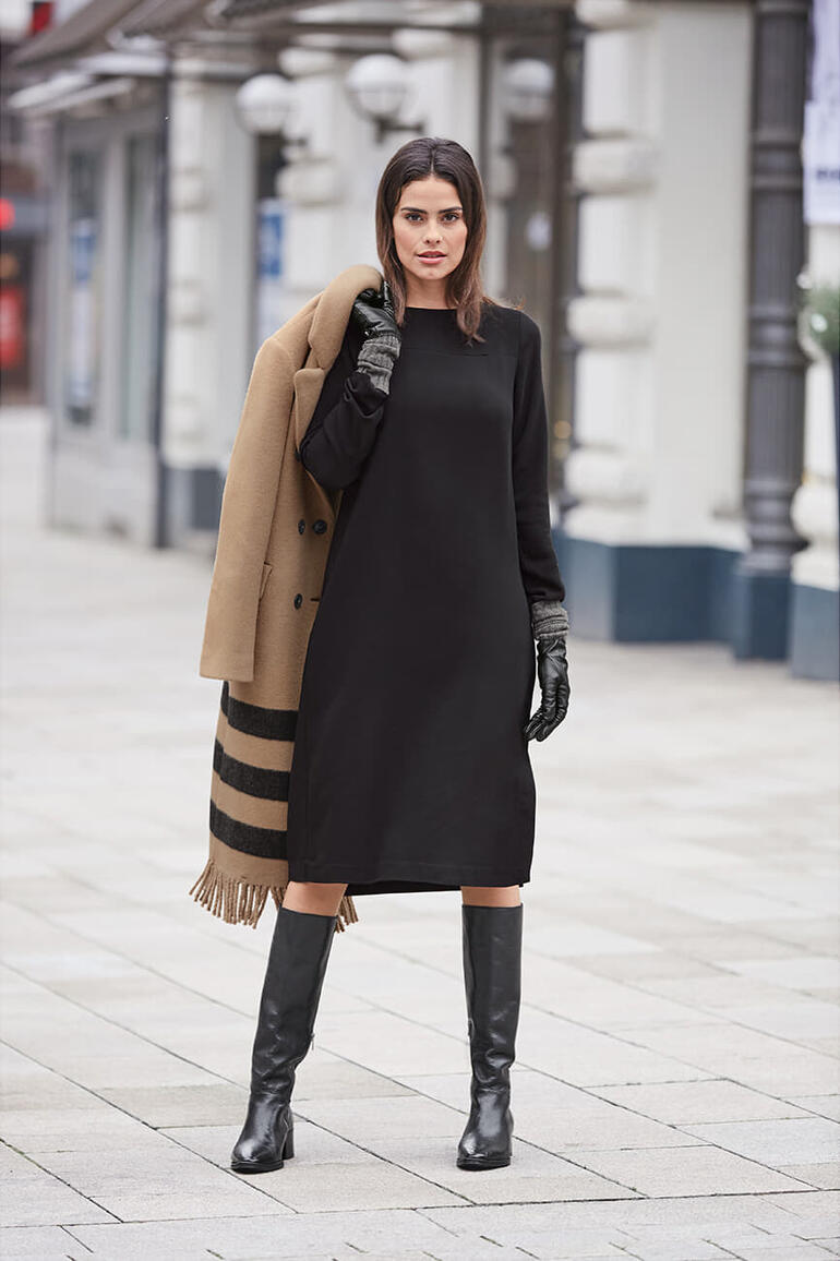 Frau in schwarzem Kleid und Mantel, mit langen Stiefeln auf einem Platz.