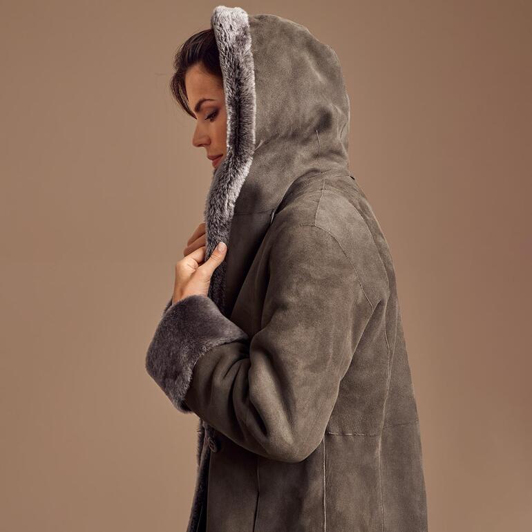 Dame im grauen Lammfell-Mantel mit Kapuze, seitliche Ansicht auf einem beigen Hintergrund.
