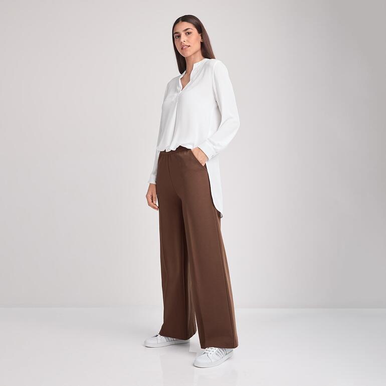 Modische braune Weitbeinformhose mit weißen Sneakers, kombiniert mit einem orangefarbenen Pullunder.