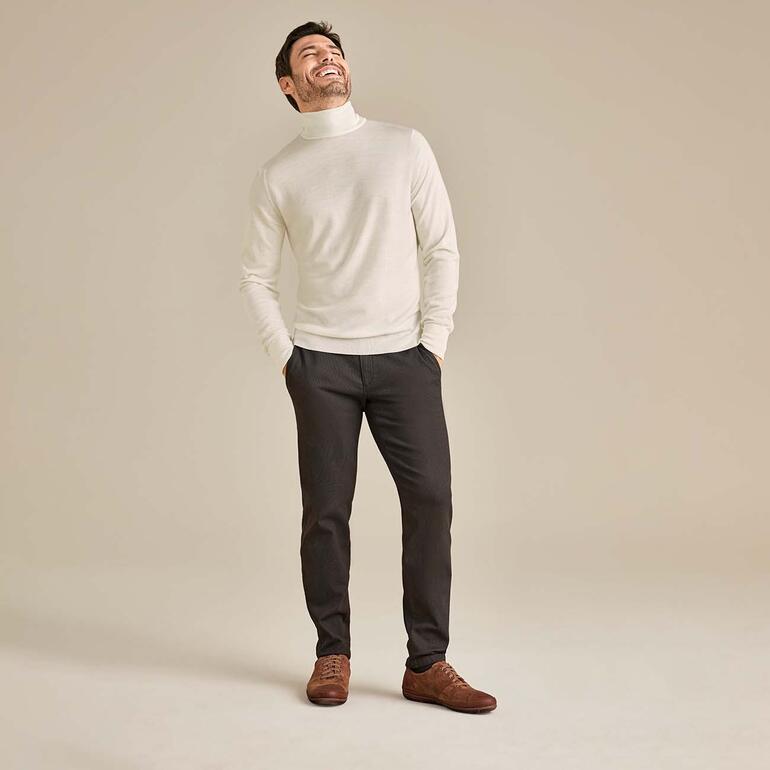 Mann in weißem Rollkragenpullover und grauen Hosen, lächelnd.