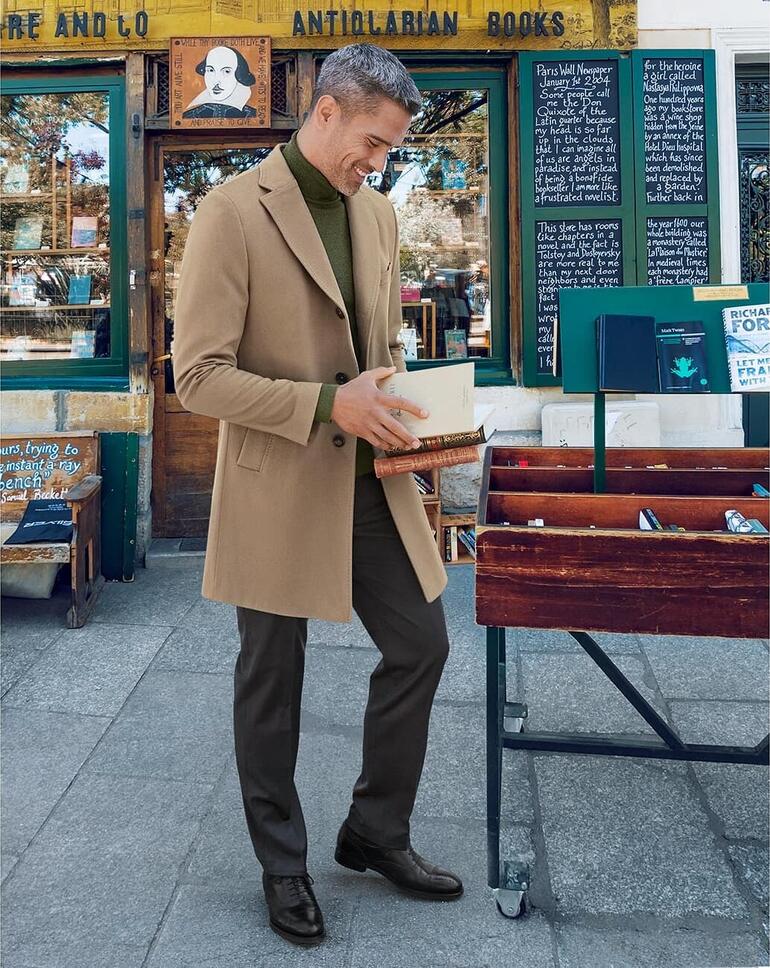 Camel Mann in Kaschmirmantel in Beige, steht vor einem Buchladen