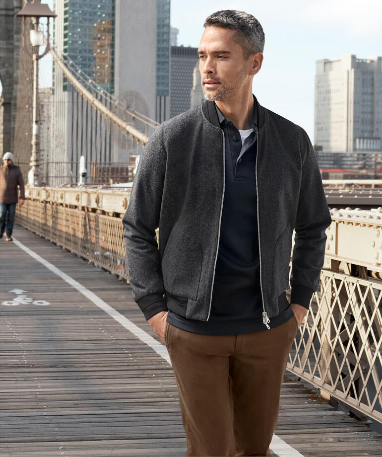 Mann in schwarzem Pima-Interlock-Poloshirt, steht in einer natürlichen Umgebung mit Bergen im Hintergrund.