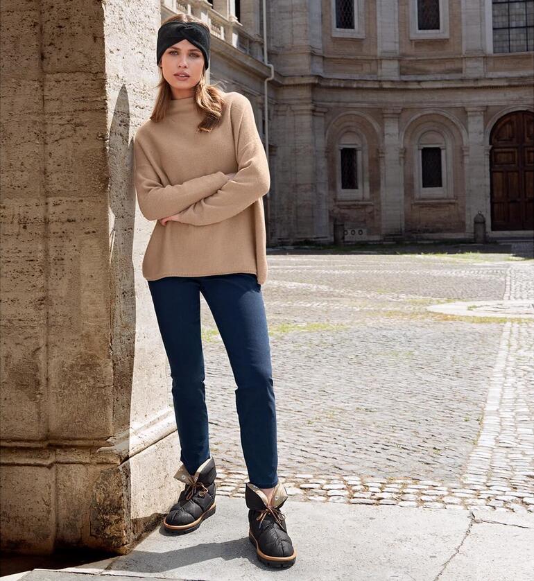 Frau in beigem Pullover und Blauen Jeans steht mit Flufié Daunen-Sockboots auf Steinboden.