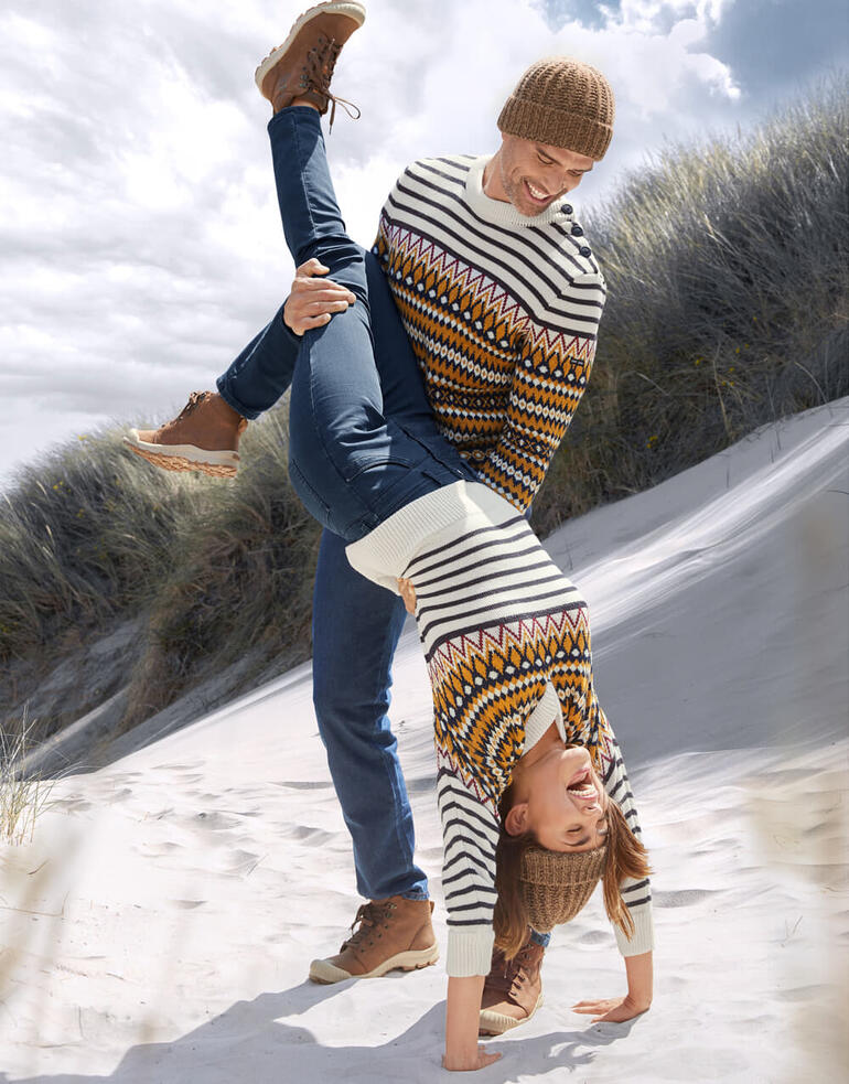 Mann hebt Frau spielerisch über den Strand, beide in Jacquard-Pullovern.