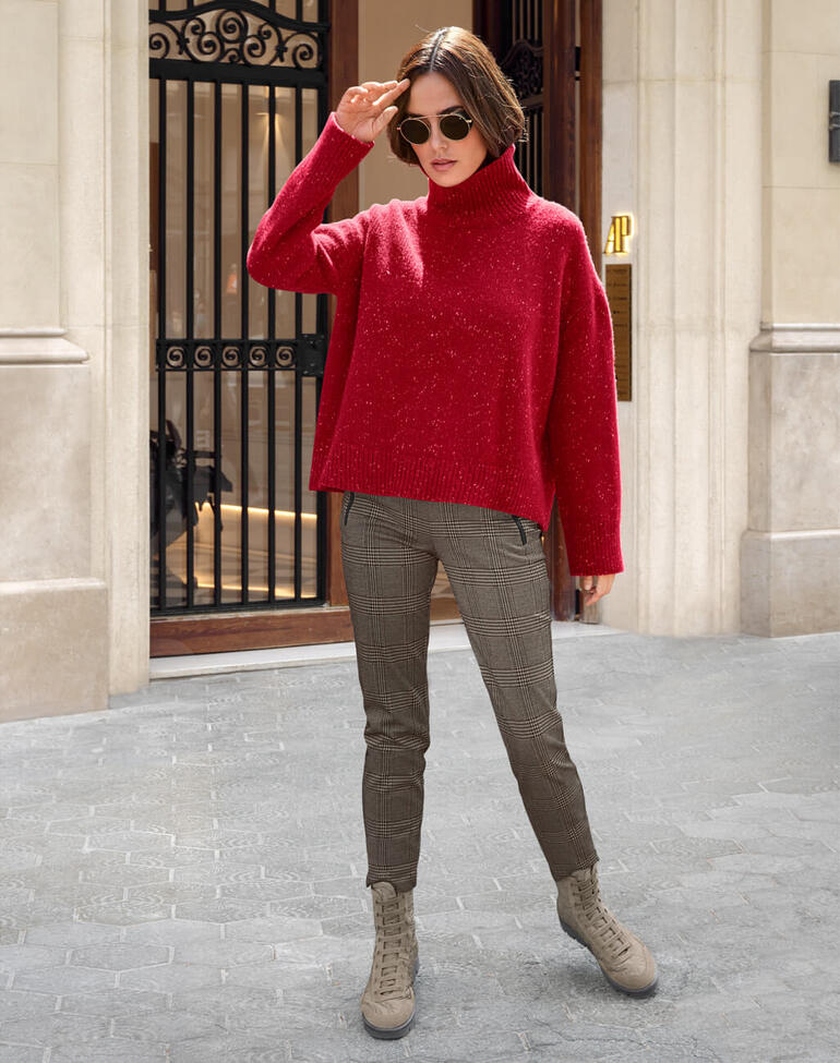 Beige Frau in rotem Pullover und karierten Schlupfhosen steht vor einer Gebäudefassade.