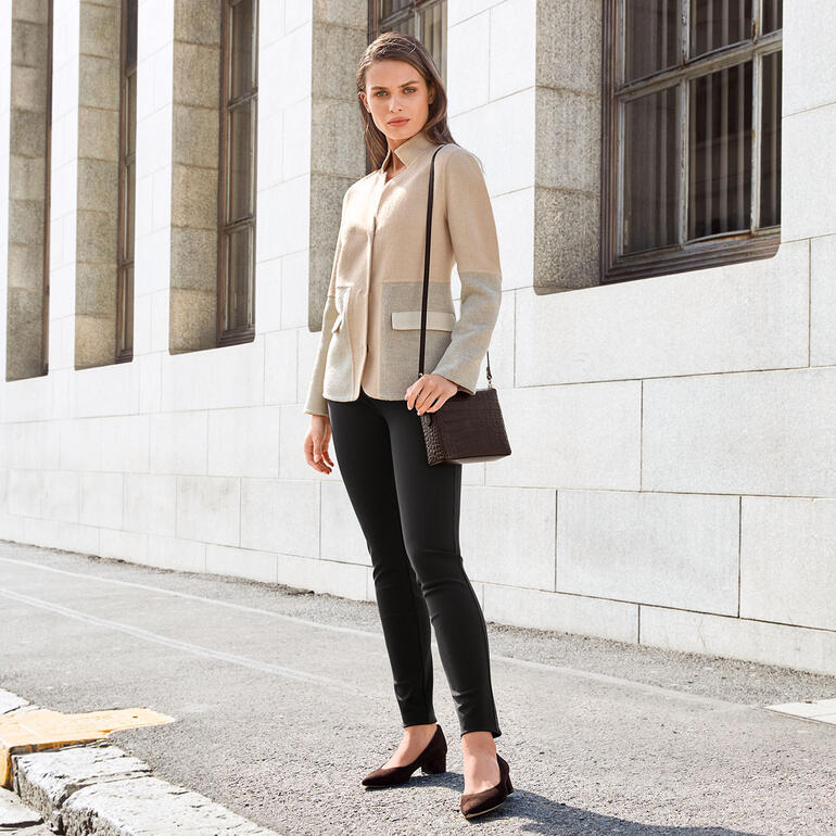 Frau in alpaka-farbigem Blazer mit Tasche, steht gegen eine Wand.