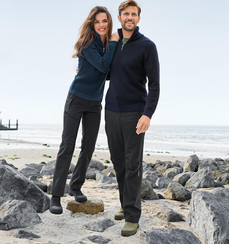 Schwarz und Beige Paar in Seemannstroyern am Strand, umgeben von Steinen und Meer.