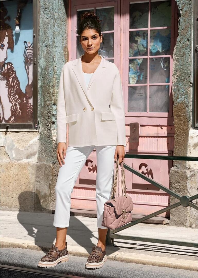 Creme Frau in hellem Blazer, braunen Hosen, sitzt auf einer Bank.