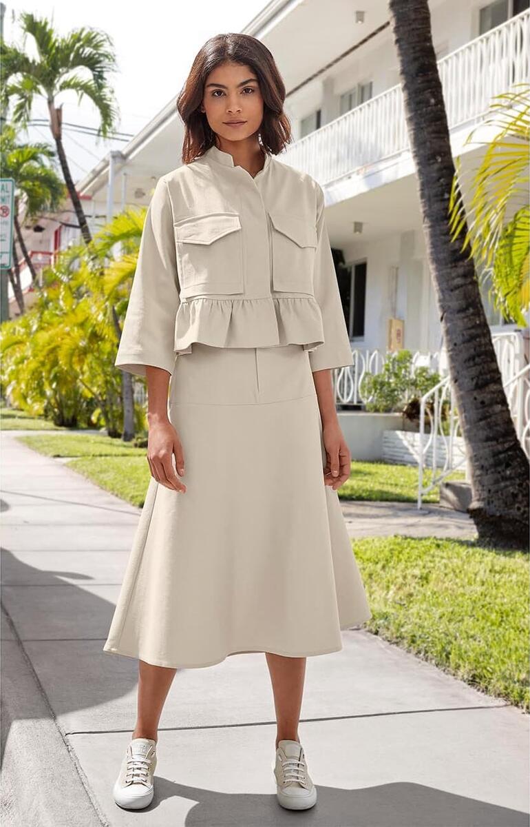 Dame in einer beige Denim-Couture-Jacke und einem A-Linien-Rock auf einer Bürgersteig mit Palmen.