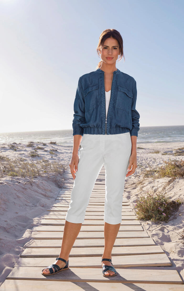 Frau im Light-Denim-Blouson und weißen Caprihosen steht am Strand.