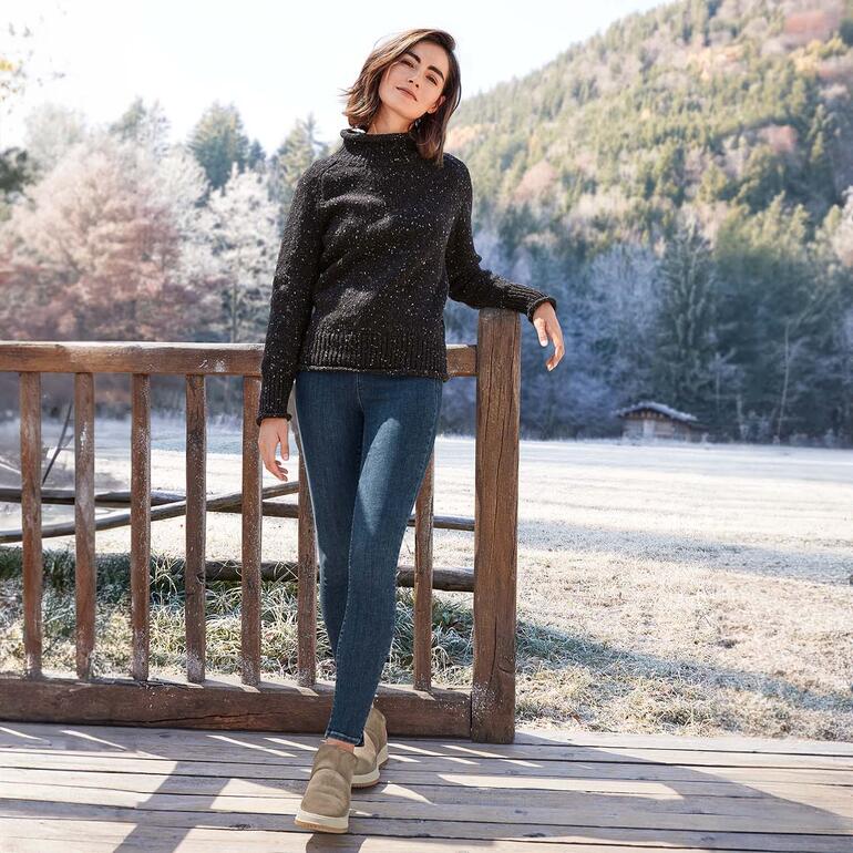 Frau in schwarzem Pullover und Jeans im Freien mit verschneiten Bäumen im Hintergrund.