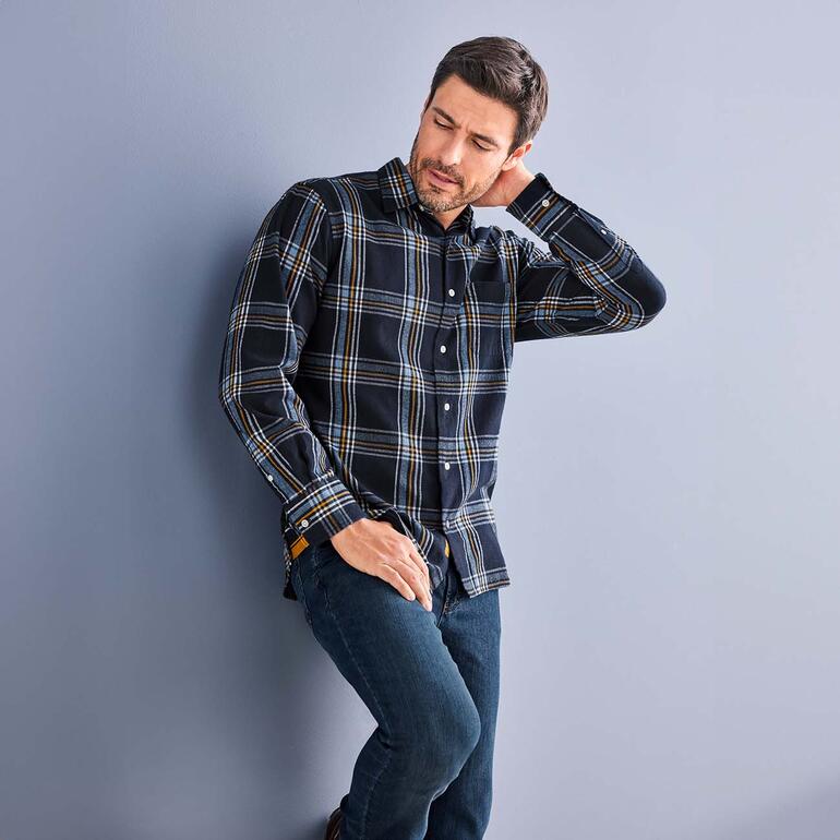 Mann in kariertem OMTC Winter-Madras-Hemd lehnt an einer Wand.