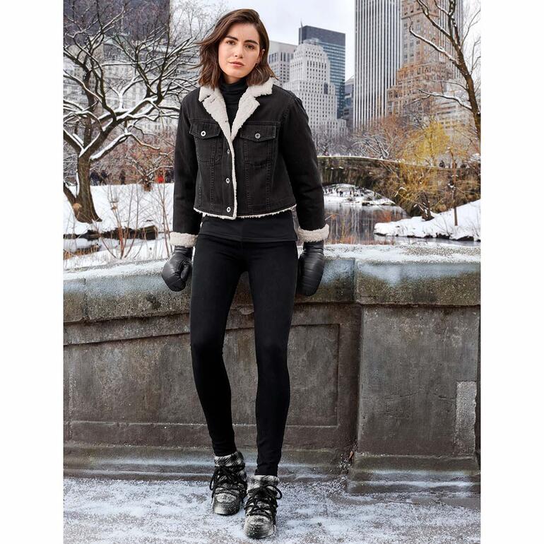 Frau in 3D shaping-Jeans mit schwarzer Jacke und warmen Handschuhen in einer winterlichen Stadtlandschaft.
