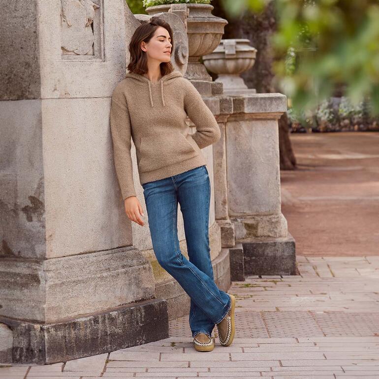Silver Suki Bootcut-Jeans Frau in Kamelhaar-Hoodie lehnt gegen eine Säule im Freien