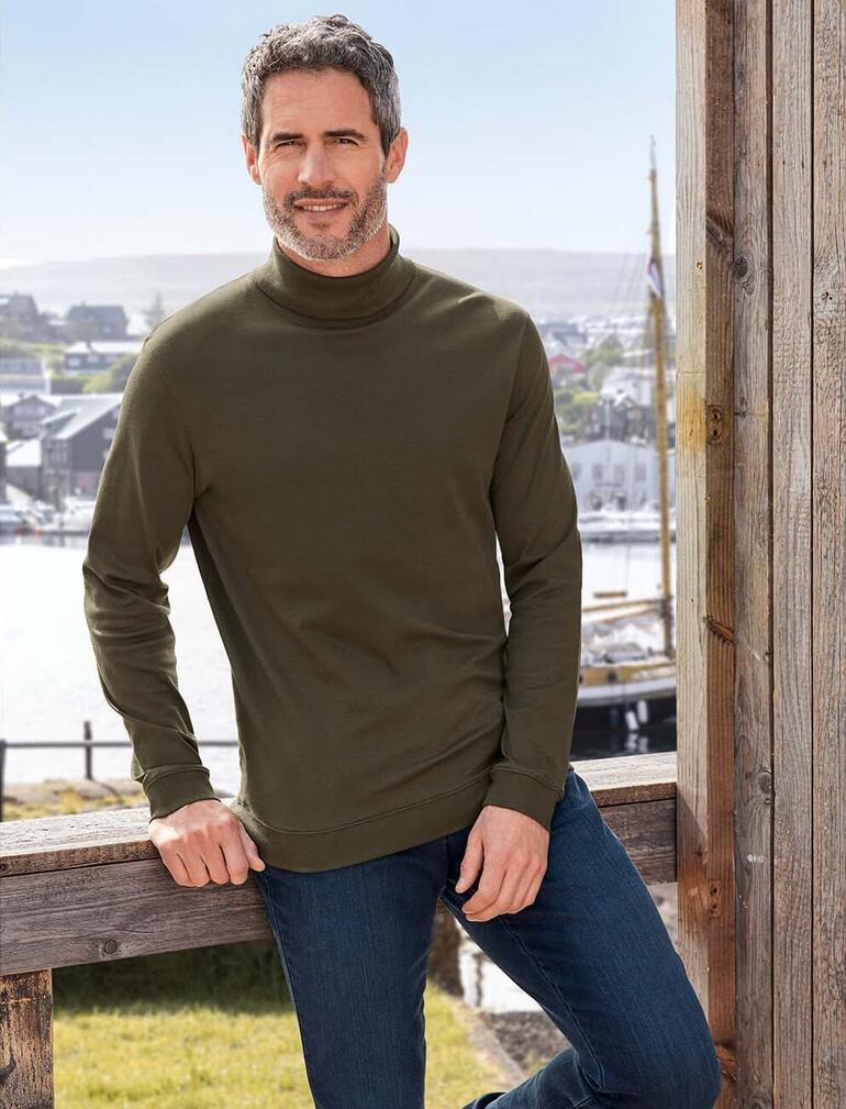 Mann trägt olivgrünen Rollkragenpullover vor einer Hafenlandschaft.
