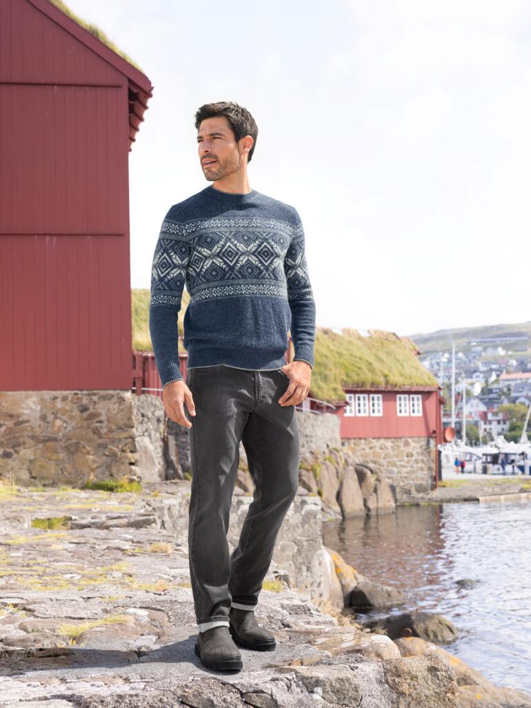 Mann in Norwegerpullover und Jeans steht am Wasser.