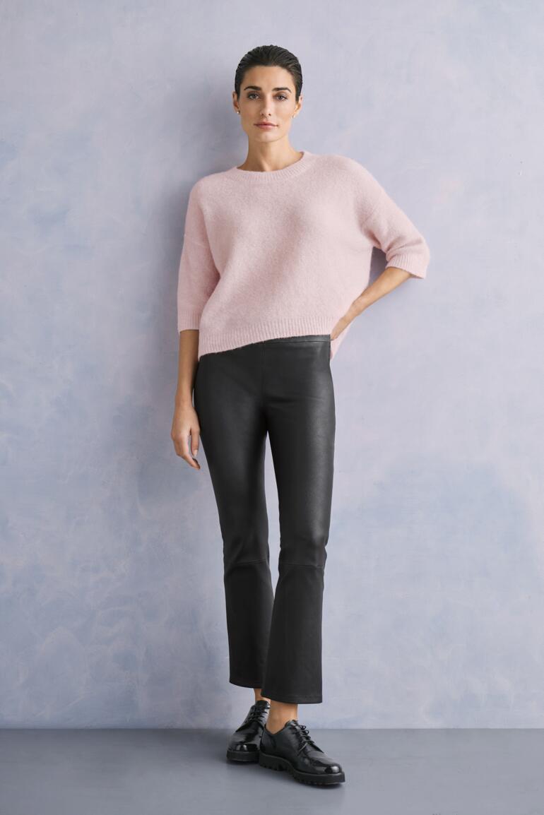 Frau in pinkem Sweater und schwarzer Kick-Flare-Lederhose vor grauem Hintergrund.