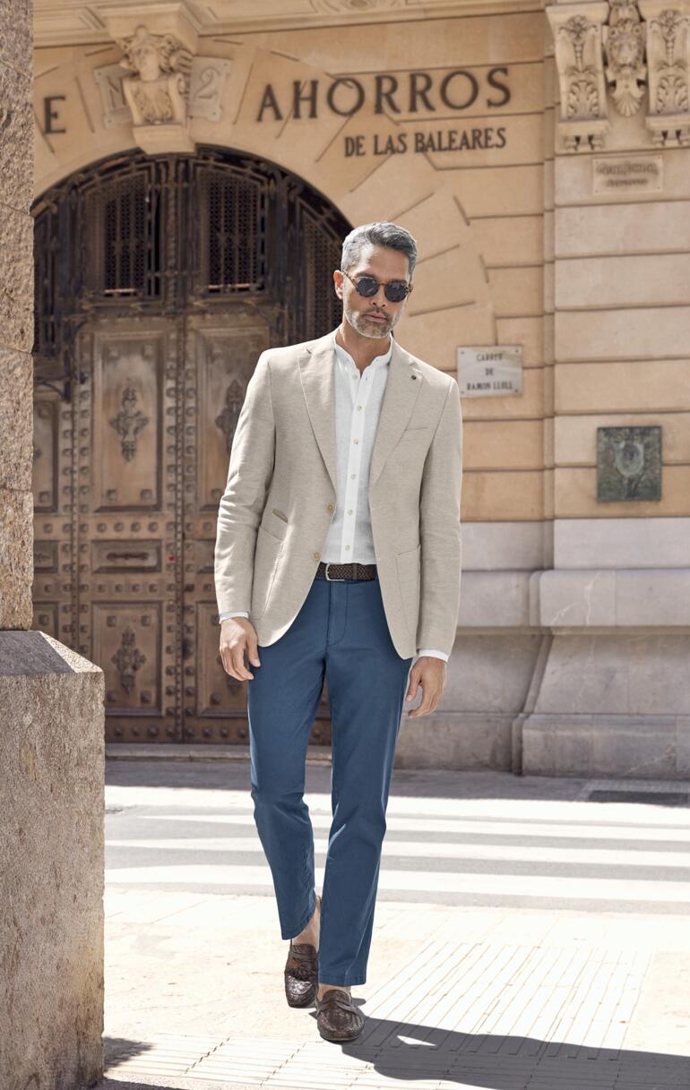 Mann in beigem Business-Leinen-Sakko, das er über blauem Hosen trägt, geht an einer historischen Kulisse vorbei.