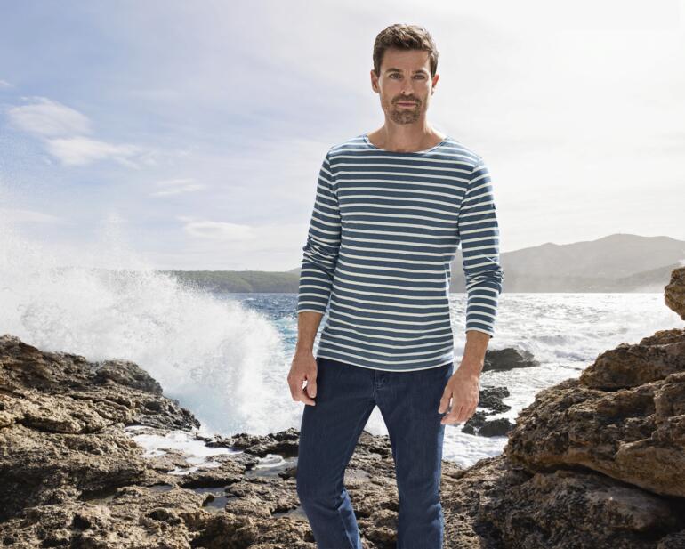 Mann trägt gestreiftes Langarmshirt und Jeans, steht an der Küste.