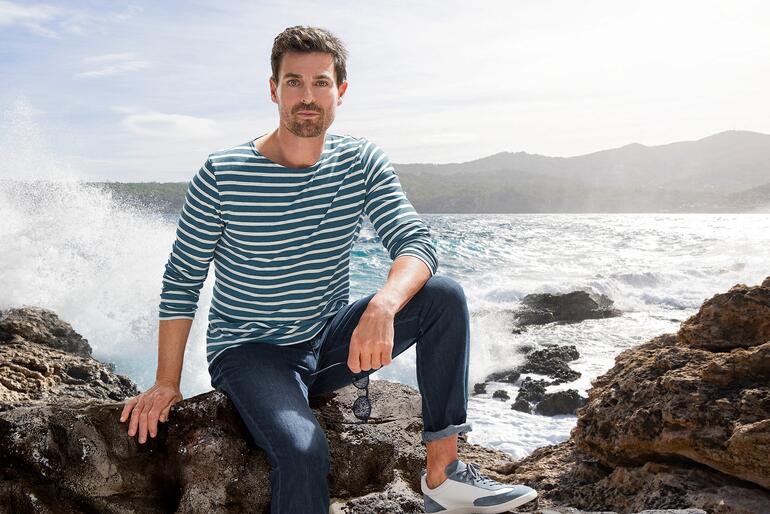 Mann im gestreiften Langarmshirt sitzt an einem Felsen am Meer.