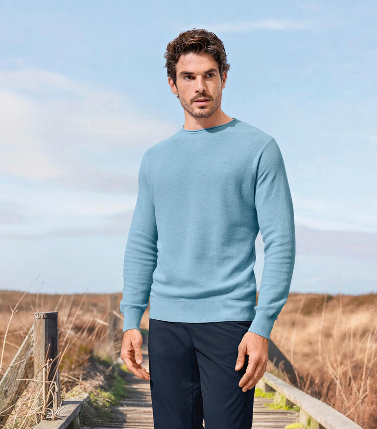 Junghans 1954 Edel-Piqué-Pullover Mann in einem hellblauen Edel-Piqué-Pullover steht in einer ländlichen Umgebung.
