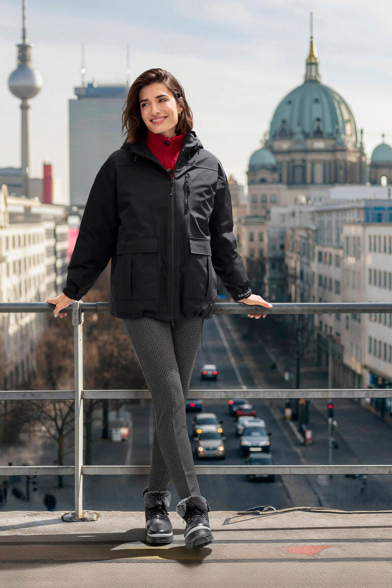 Frau in schwarzer Daunen-Regenjacke vor städtischer Kulisse.