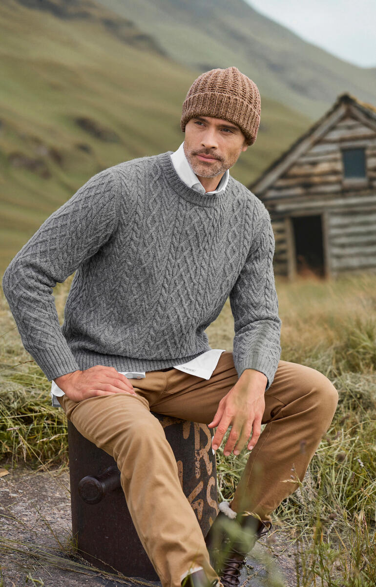 Mann sitzt auf einem Stein, trägt einen grauen Aran-Pullover und Mütze.