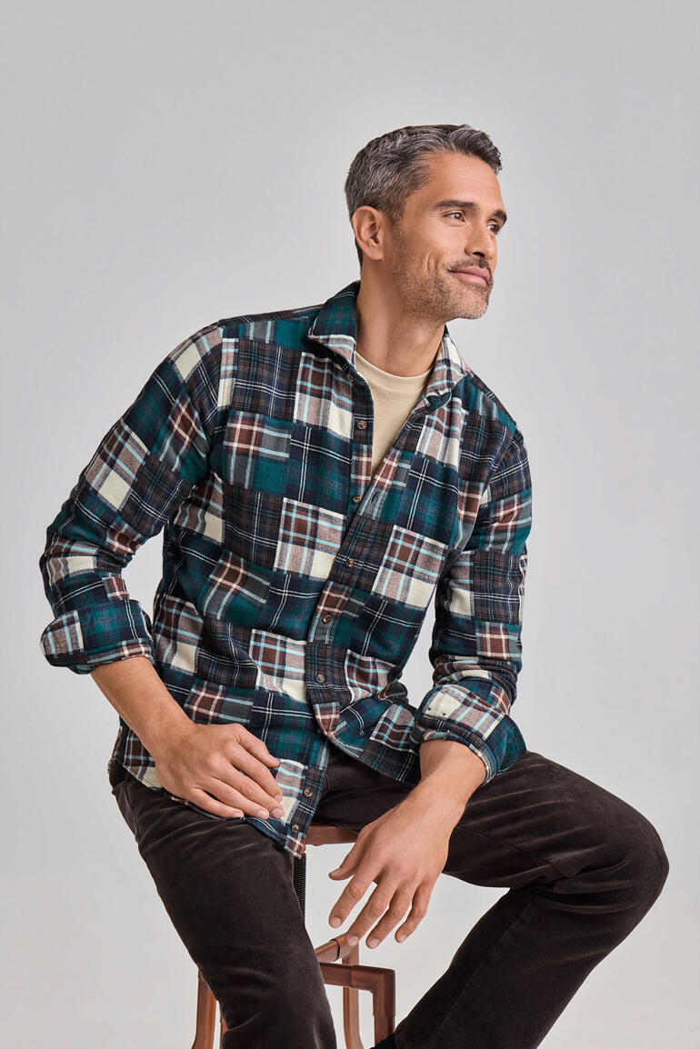 Mann trägt ein DU4 Patchwork-Flanellhemd mit Karomuster, sitzt auf einem Hocker.
