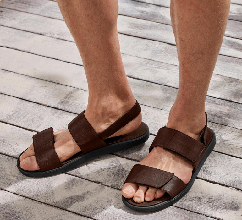 Ultralight-Ledersandale Braunes Leder-Sandale mit zwei Riemen an einem Fuß auf einem Holzfußboden.