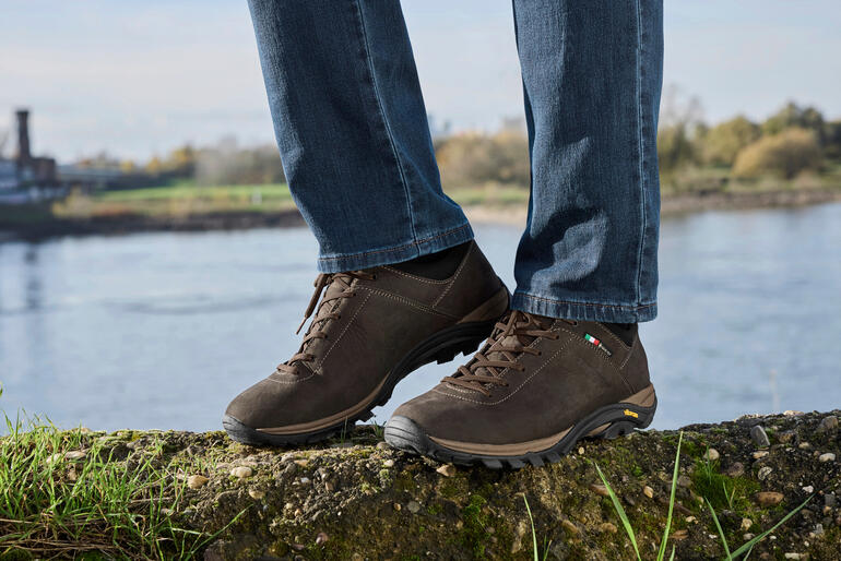 Herren, Dunkelbraun Braune Zamberlan Sneaker Commute GTX auf einem Felsen nahe einem Gewässer.