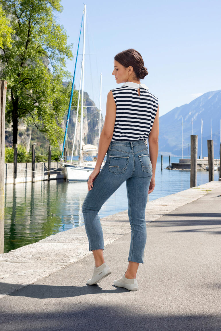 Frau in gestreiftem Oberteil und Jeans auf einem Gehweg am Wasser.
