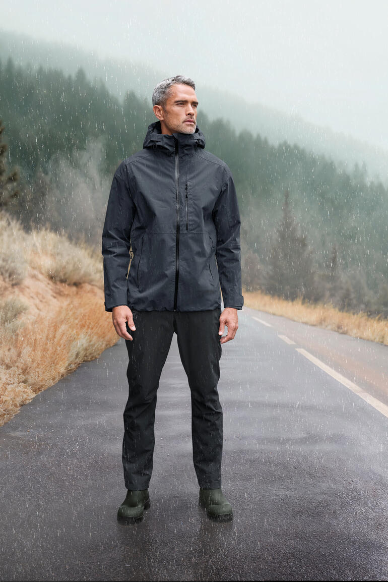 Mann in grauer Hardshell-Jacke steht auf nasser Straße in bewaldeter Landschaft.