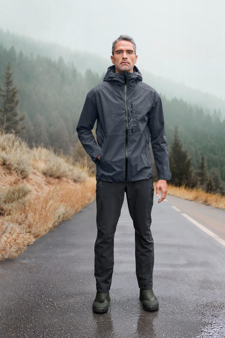 Mann in grauer Hardshell-Jacke steht auf nasser Straße, im Hintergrund Berge.