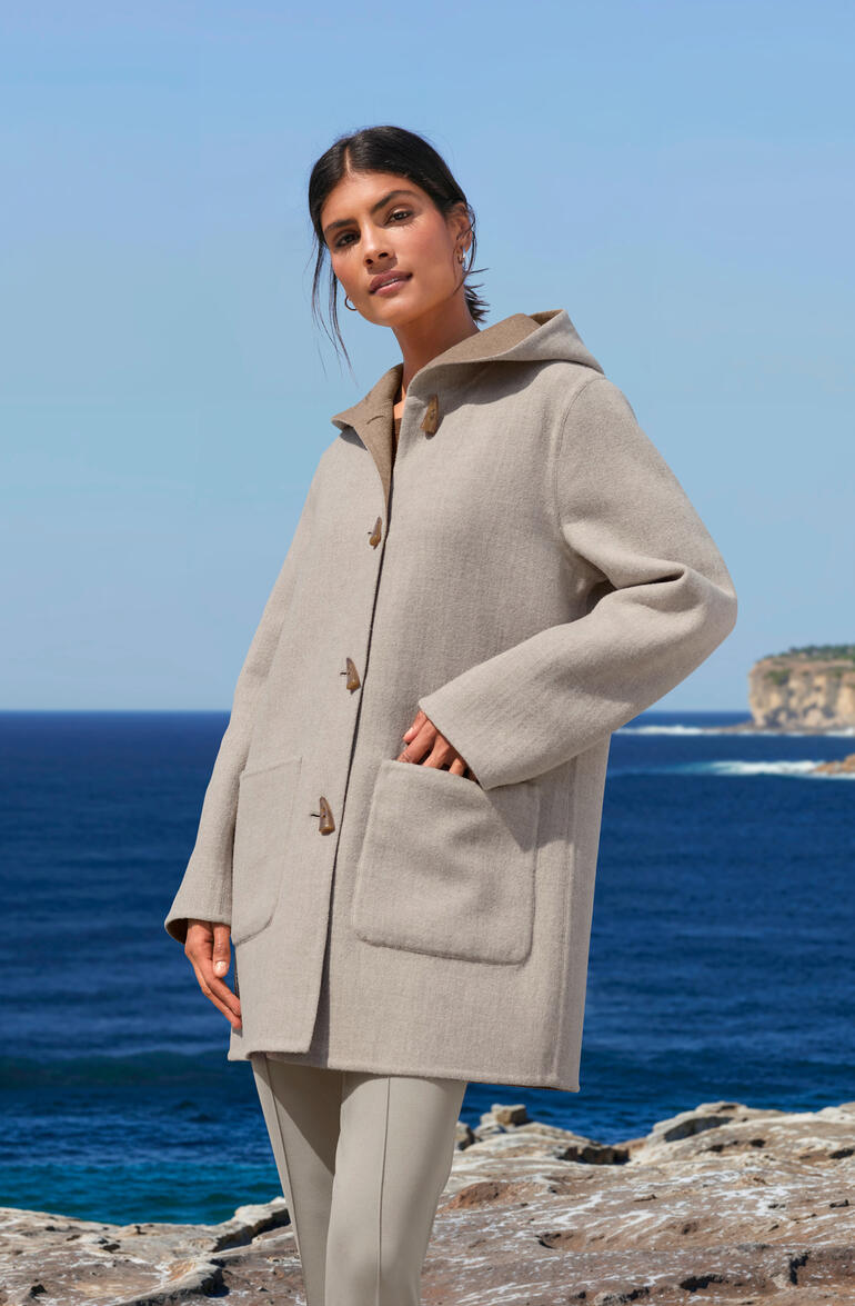 Alpaka-Doubleface-Dufflecoat in Beige, getragen von einer Frau mit Meer im Hintergrund.