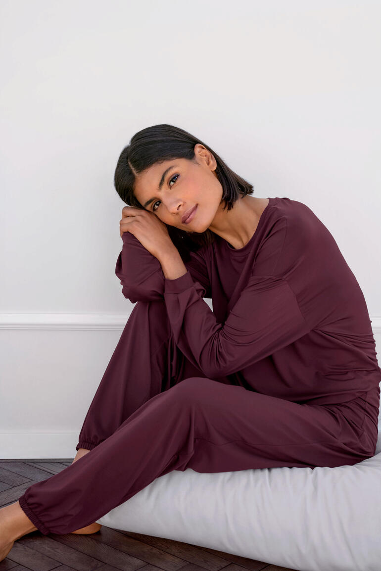 Frau sitzt in einem lila Loungewear Shirt und Hose auf einem Kissen.