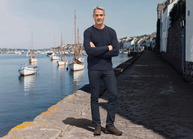 Mann in einem dunkelblauen Rippenpullover, steht am Hafen mit Booten im Hintergrund.