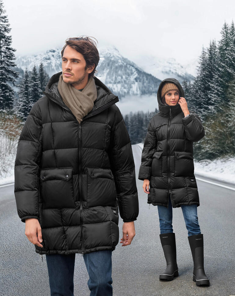 Zwei Personen in schwarzen Daunen-Parkas auf einem winterlichen Straßenabschnitt mit Bergen im Hintergrund.