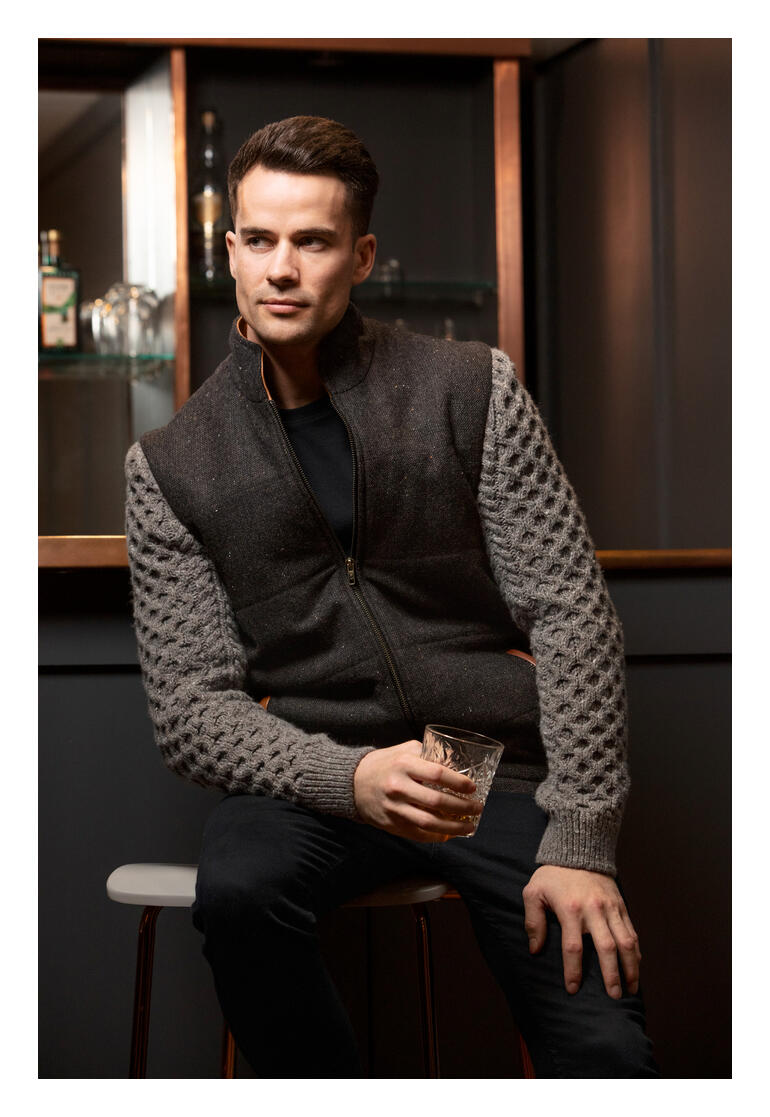 Celtic Tweed Hybrid-Jacke Mann trägt eine braun-graue Celtic-Tweed-Jacke, sitzt mit einem Glas.