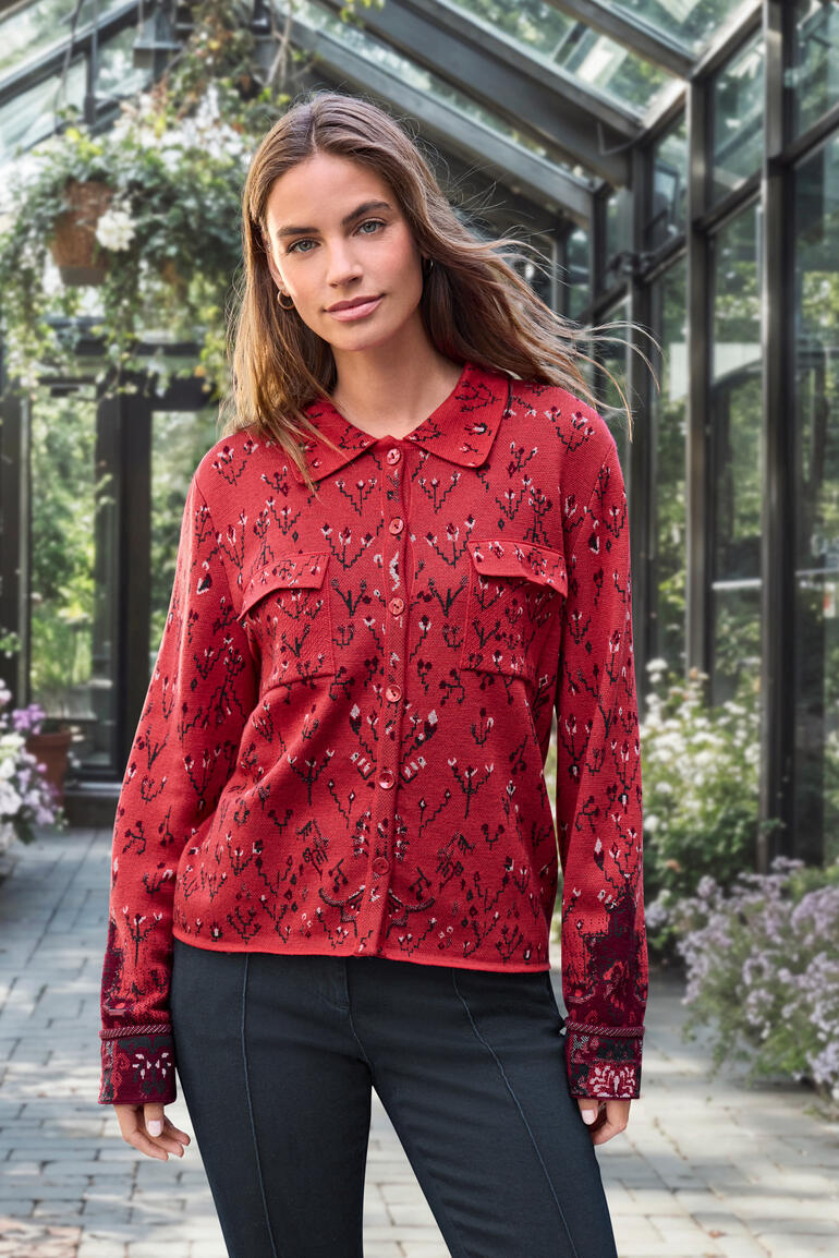 Frau in roter Jacquard-Strickbluse mit floralem Muster in einem Gewächshaus.