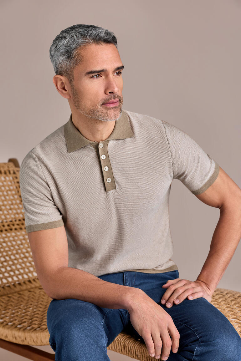 Mann trägt sand-beiges Polohemd mit Knöpfen und sitzt auf einem Stuhl.
