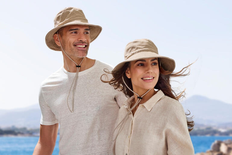 Pärchen trägt sandfarbene Strandhüte am Meer, lächelnd zur Kamera.