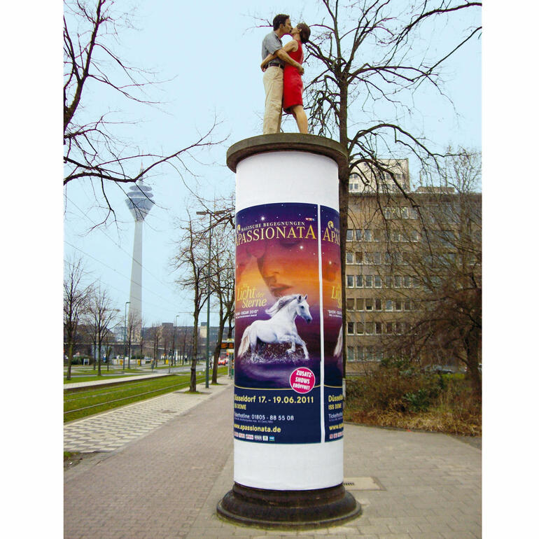 Die 1,90 m grosse Originalskulptur auf einer Litfass-Säule ist in der Düsseldorfer Innenstadt (Ecke Haroldstrasse/Poststrasse) zu sehen. Paar, das sich auf einer Säule in der Stadt umarmt, umgeben von Werbeplakaten.