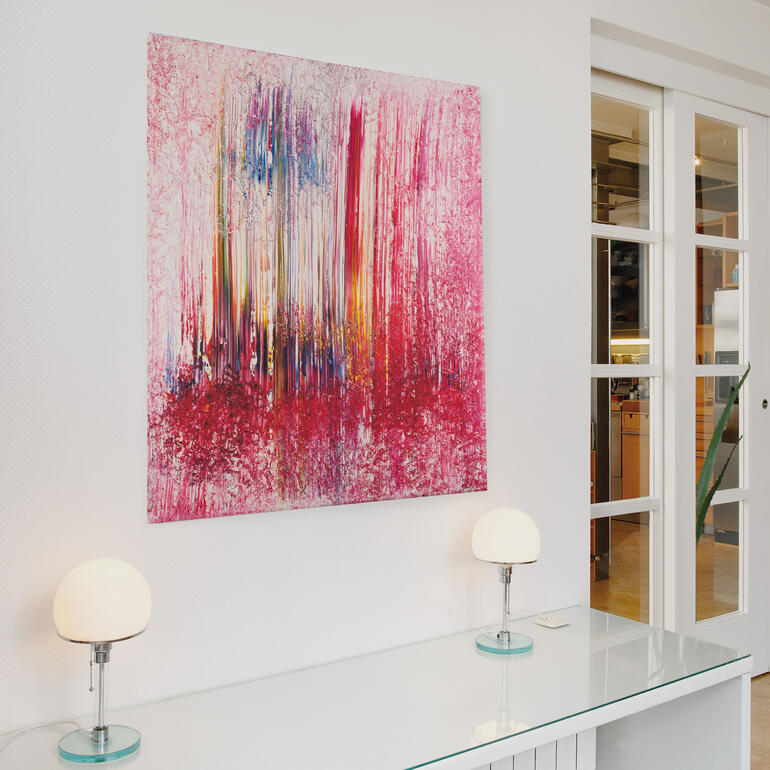 Abstraktes Wandbild in Pink- und Rottönen, hängt über einem modernen Tisch mit Lampen.